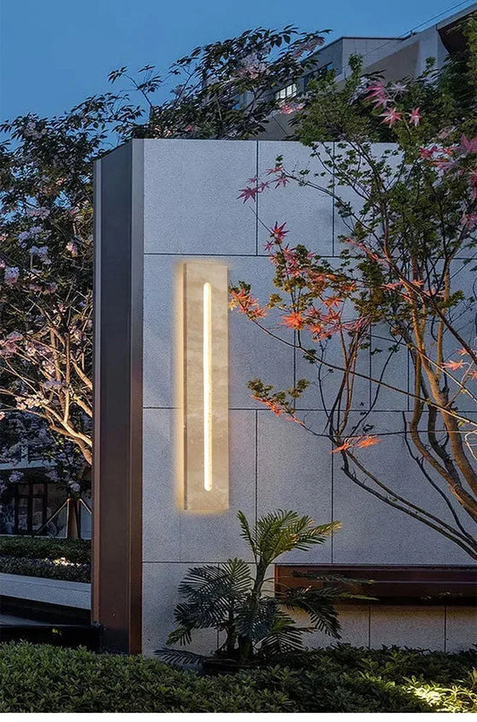Rectangular wall sconce with a natural stone finish and a vertical LED light, mounted on a textured gray wall outdoors at dusk.