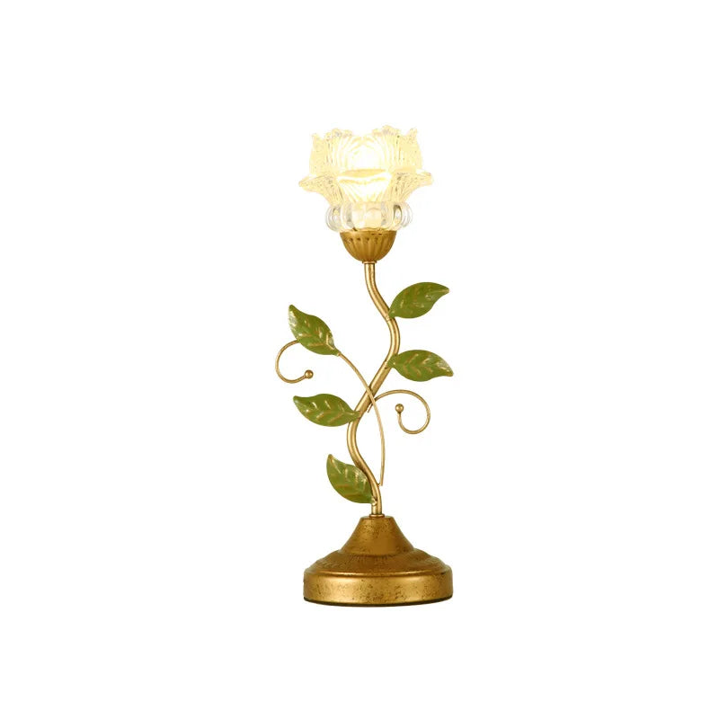 A white background product shot of a gold table lamp shaped like a flower with a glass shade and green leaves.