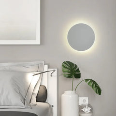 A white circular wall sconce mounted next to a bed with a plant in a vase.