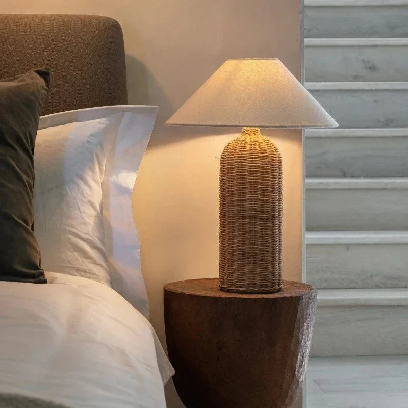 Woven rattan table lamp with a fabric shade placed on a wooden bedside table next to a bed.