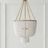 A close-up studio shot of a gold-framed chandelier adorned with numerous white beads, hanging from a gold chain against a white paneled wall.
