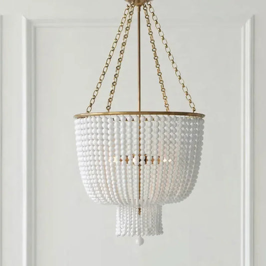 A close-up studio shot of a gold-framed chandelier adorned with numerous white beads, hanging from a gold chain against a white paneled wall.