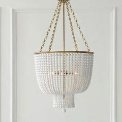 A close-up studio shot of a gold-framed chandelier adorned with numerous white beads, hanging from a gold chain against a white paneled wall.