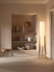 A floor lamp with a black walnut tripod base and a white fabric shade, illuminated in a room with built-in shelves displaying decorative items.