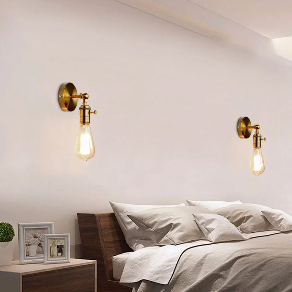 A bedroom setting with two gold industrial-style wall sconces, each with an adjustable folding arm and exposed Edison-style bulb, mounted on the wall above a bed.