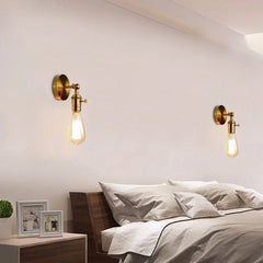 A bedroom setting with two gold industrial-style wall sconces, each with an adjustable folding arm and exposed Edison-style bulb, mounted on the wall above a bed.