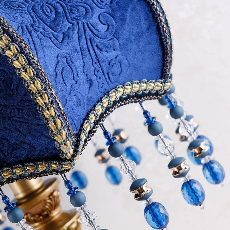 Close-up of the blue fabric shade of a table lamp, showing the textured pattern, gold trim, and various crystal and blue bead strands hanging down.