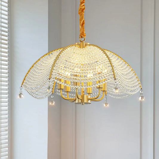 A close-up, front-facing view of a gold-finish chandelier with a fan-shaped crystal-covered canopy and hanging crystal teardrops, illuminated against a softly lit wall.
