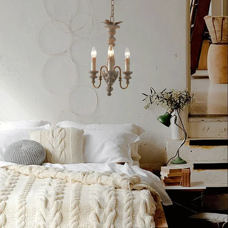 Distressed wood candelabra pendant light with three exposed bulbs installed above a bed in a rustic bedroom setting with white bedding and cable knit throws.