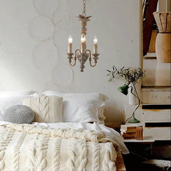Distressed wood candelabra pendant light with three exposed bulbs installed above a bed in a rustic bedroom setting with white bedding and cable knit throws.