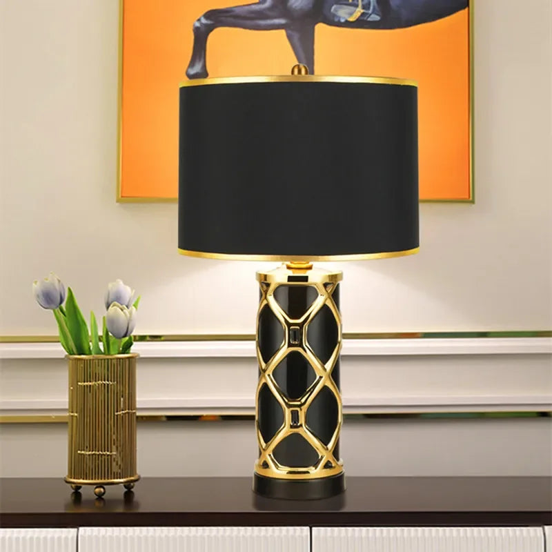 Decorative table lamp with black and gold design on a surface, with a vase of flowers and abstract art in the background.