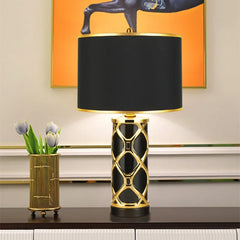 Decorative table lamp with black and gold design on a surface, with a vase of flowers and abstract art in the background.