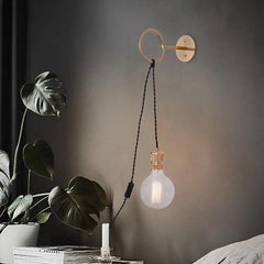 A gold ring wall sconce with a clear glass bulb hanging from a black cord, styled in a bedroom setting with plants.