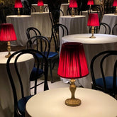 A close-up of a burgundy pleated fabric shade table lamp with a gold base, illuminated from within, sitting on a white tabletop.