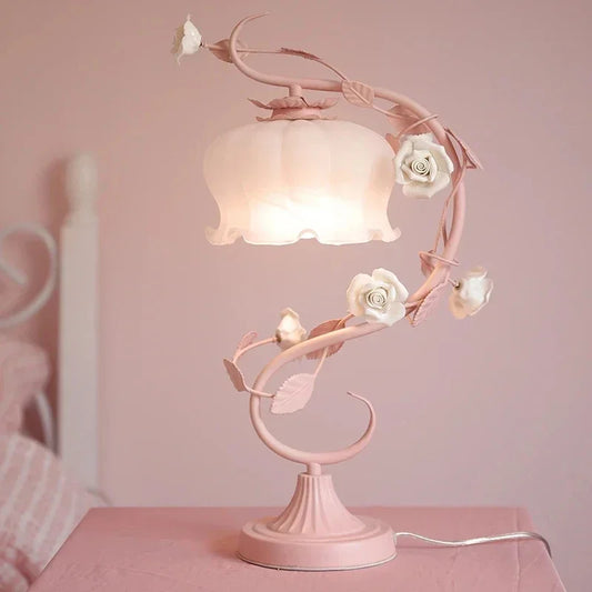 A pink table lamp with a floral rose vine design and a white frosted bell-shaped shade, illuminated on a pink surface.