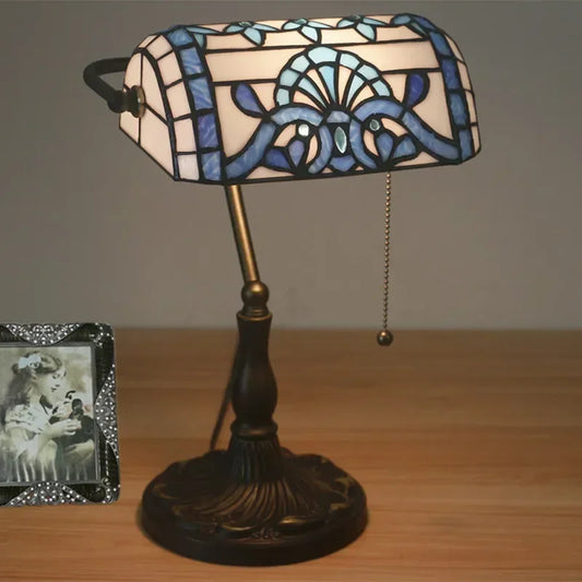 A Tiffany-style stained glass table lamp with a blue and white patterned shade, a bronze finish base, and a pull chain, placed on a wooden surface next to a picture frame.