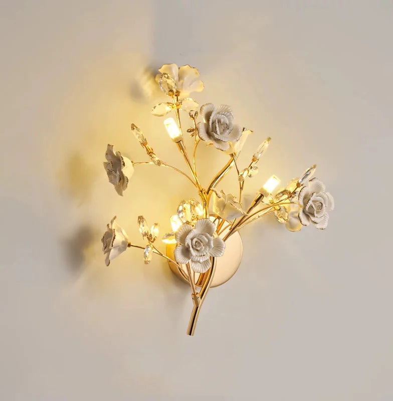 A gold wall sconce with white ceramic flowers and clear crystals, emitting a warm light.
