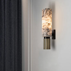 A wall sconce with a natural marble shade and reeded brass base mounted on a wall, with a dark gray curtain to the left.