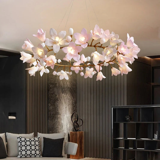 A circular chandelier with pink and white flower-shaped lights and gold branches, illuminated and hanging in a modern living room.