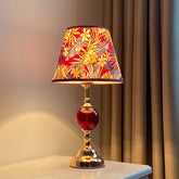 A polished brass table lamp with a conical red floral fabric shade and a round red glass orb accent, shown on a white surface.