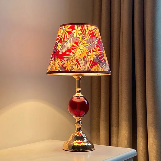 A polished brass table lamp with a conical red floral fabric shade and a round red glass orb accent, shown on a white surface.