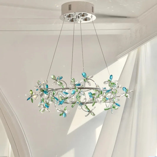 Close-up of a chrome ring chandelier adorned with clear and blue/green crystal flowers, suspended by thin wires against a white ceiling.