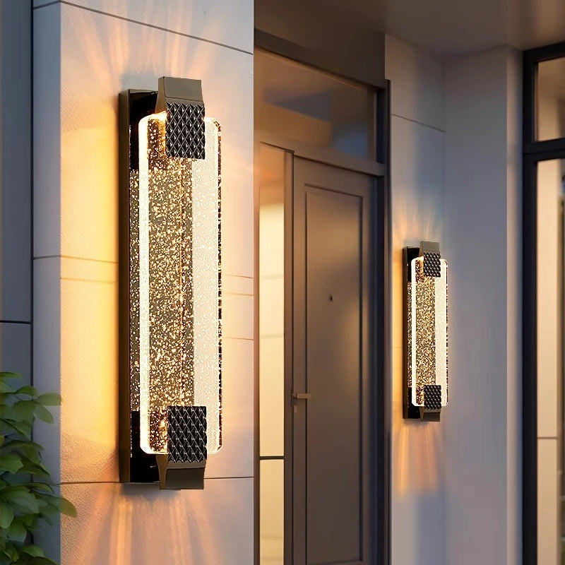A gold wall sconce with a crystal diffuser illuminated on a marble column next to an entryway.