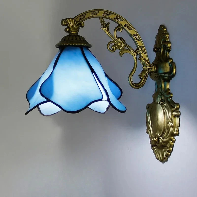 A wall-mounted light fixture with an antique gold metal arm and a scalloped blue stained glass shade, emitting a soft light.