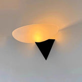 A modern wall sconce with a frosted oval shade mounted on a black triangular base, casting a warm light on a white wall.