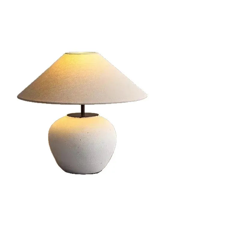 A white ceramic pot-shaped table lamp with a conical fabric lampshade is shown against a white background.
