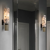 A wall sconce with a cylindrical natural marble shade and a reeded brass base, mounted on a gray wall next to a mirror.