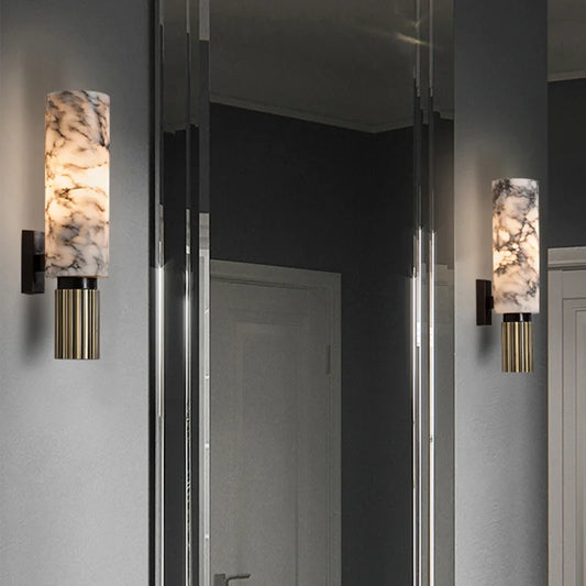 A wall sconce with a cylindrical natural marble shade and a reeded brass base, mounted on a gray wall next to a mirror.