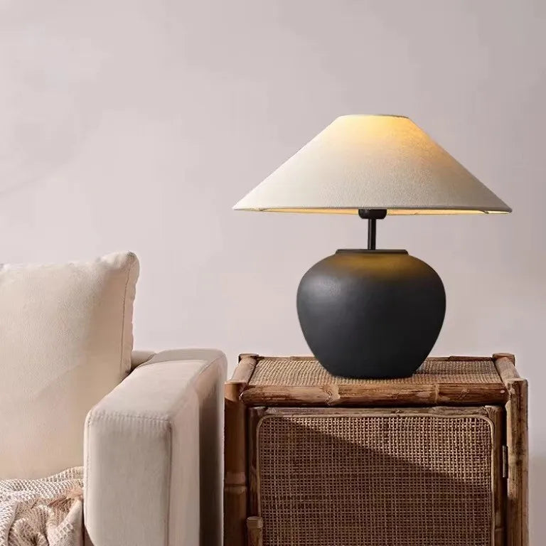 A matte black ceramic pot-shaped table lamp with a conical fabric lampshade is placed on a small wooden side table next to a sofa.