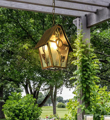 Decorative outdoor light fixture hanging from a pergola with greenery in the background