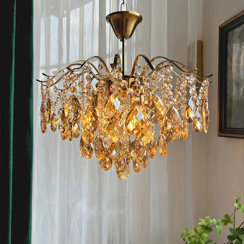 Gold chandelier with crystal accents hanging in a room with curtains and a plant.