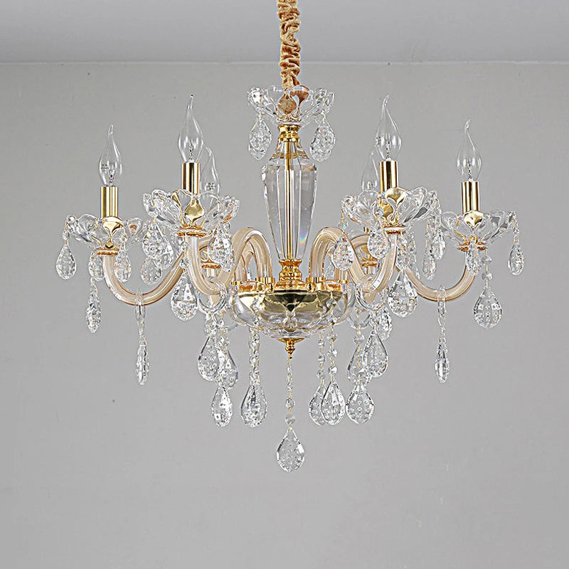 A gold crystal chandelier with multiple arms and dangling crystals, shown in a close-up view against a light gray wall.