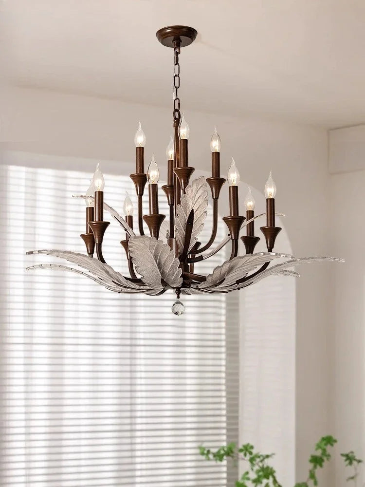 A wide shot of a large bronze chandelier with metal leaf details and candle-style bulbs, illuminating a room with natural light from windows.