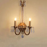 A close-up view of a gold wall sconce with two candle-style lights and dangling clear crystals against a light yellow wall.