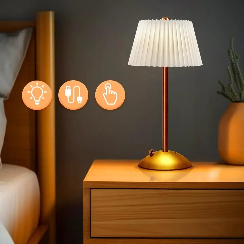 A white pleated fabric shade table lamp with a gold base and stem, placed on a wooden nightstand next to a bed, with icons indicating light, USB, and touch control.