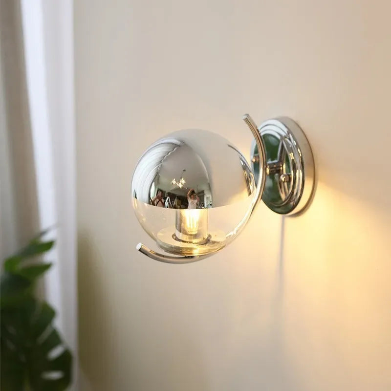 Chrome wall-mounted light fixture on a neutral wall with a plant in the corner.