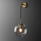 A gold wall sconce with a clear glass globe hanging from an arm mounted on a dark gray wall.