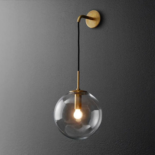 A gold wall sconce with a clear glass globe hanging from an arm mounted on a dark gray wall.