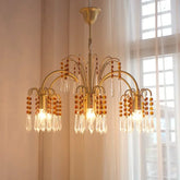 A gold-toned chandelier with cascading crystal and amber beads, illuminated against a backdrop of sheer white curtains.