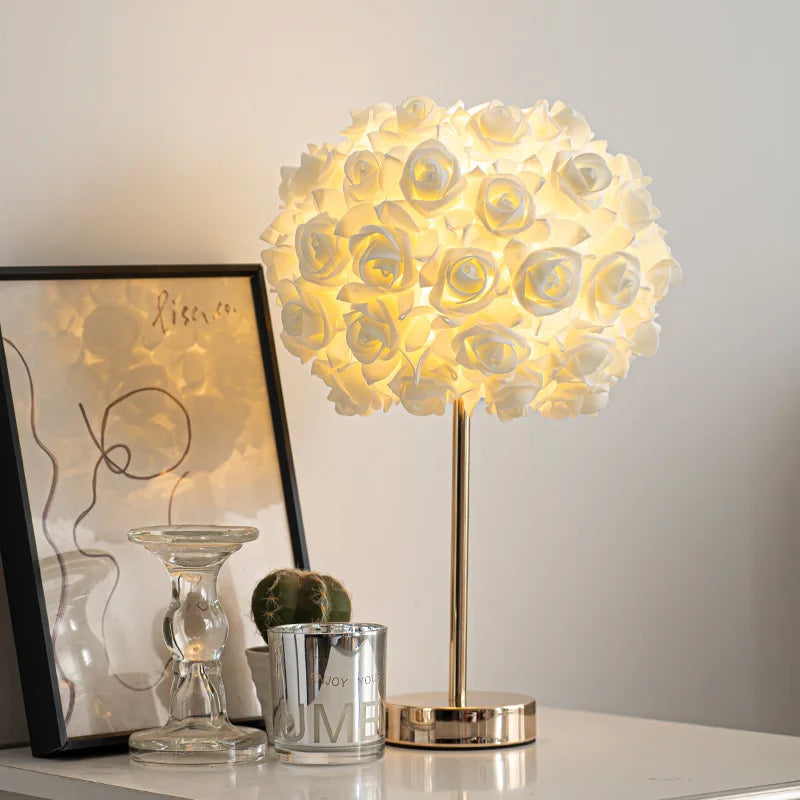 A white floral rose bud table lamp with a gold base and stem, placed on a desk next to a framed picture and a candle.