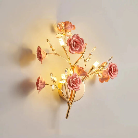 A gold wall sconce with pink ceramic flowers and clear crystals, emitting a warm light.