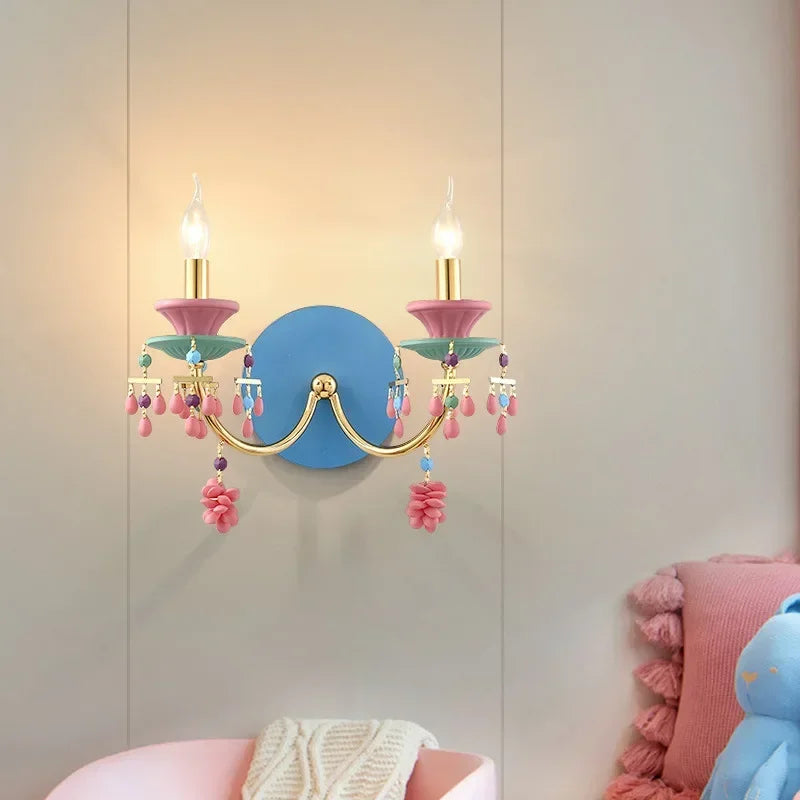 Gold wall sconce with illuminated candle-style bulbs and colorful beaded and flower-like accents, mounted on a blue backing plate, in a room setting.