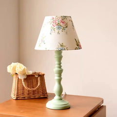 Floral-patterned lampshade on a green lamp base with a basket of flowers on a wooden surface.