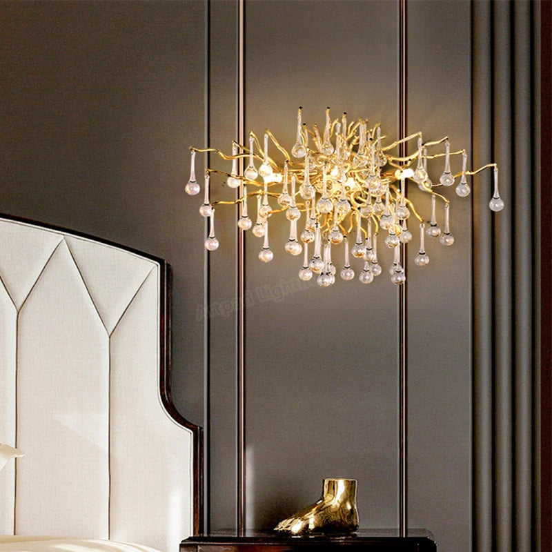 A gold crystal wall sconce with a branch-like design and teardrop crystals mounted on a dark wall next to a bed.