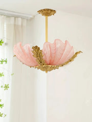 Pink seashell glass pendant light with ornate gold leaf accents and a central brass stem, hanging against a soft white curtain.