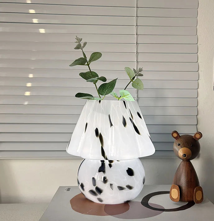 White lamp with black spots on a box next to a wooden bear toy, with a blurred window in the background.